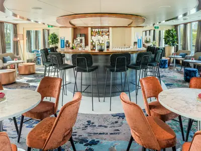 Innenansicht der Lounge-Bar auf der Seine Comtesse. Eine zentrale Rundtheke mit Barhockern, orangefarbene Sessel und Tische sind sichtbar. Große Fenster bieten Ausblick auf die Flusslandschaft.
