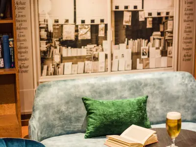 Eine gemütliche Sitzecke mit einem hellblauen Samtsofa, einem grünen Kissen und einem Bücherregal voller Bücher. Im Hintergrund ist eine große Wandmalerei eines Buchladen-Fensters zu sehen. Auf dem Tisch steht ein Bierglas.