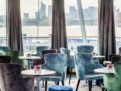 Innenansicht der Seine Comtesse Lounge. Komfortable Sessel und Tische stehen vor großen Fenstern mit Blick auf einen Fluss und die Stadtsilhouette. Eine einladende Atmosphäre.