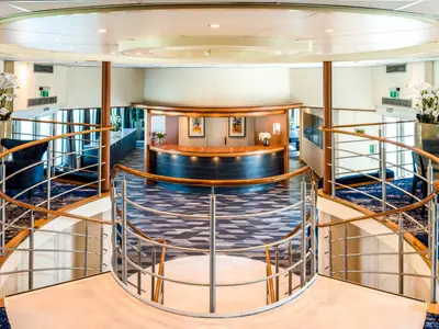 Innenbereich der Seine Comtesse, mit geschwungener Treppe, die zu einem Rezeptionsbereich mit Bar und Lounge führt. Moderner Teppich und helle Gestaltung sind zu sehen.