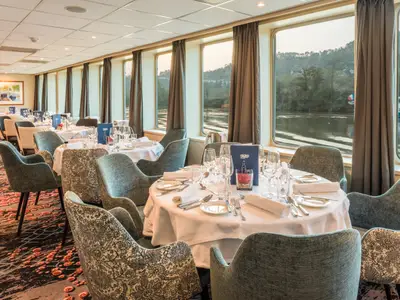 Blick in den hellen Restaurantbereich der Seine Comtesse mit elegant gedeckten Tischen und bequemen Stühlen. Durch die großen Fenster ist ein Fluss mit bewaldeten Ufern zu sehen.