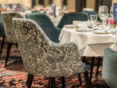 Detaillierte Ansicht eines gemusterten Stuhls mit Blumenmuster im Restaurant der Seine Comtesse. Ein gedeckter Tisch mit weißer Tischdecke, Gläsern und Besteck ist ebenfalls zu sehen.