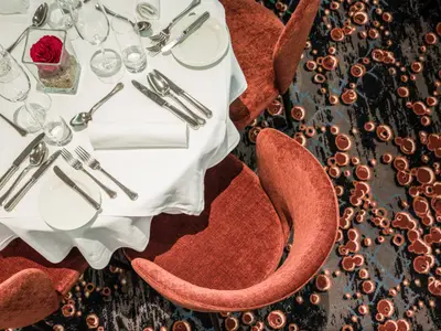 Vogelperspektive auf einen festlich gedeckten Restauranttisch mit weißer Tischdecke, Besteck, Gläsern und einer roten Rose. Zwei moderne, rostfarbene Stühle und ein gemusterter Teppich sind sichtbar.