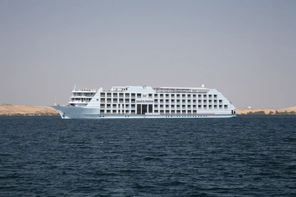 Seitenansicht des weißen Flusskreuzfahrtschiffs Steigenberger Omar El Khayam auf einem See oder Fluss, mit sandiger Uferlandschaft im Hintergrund unter einem hellen Himmel.