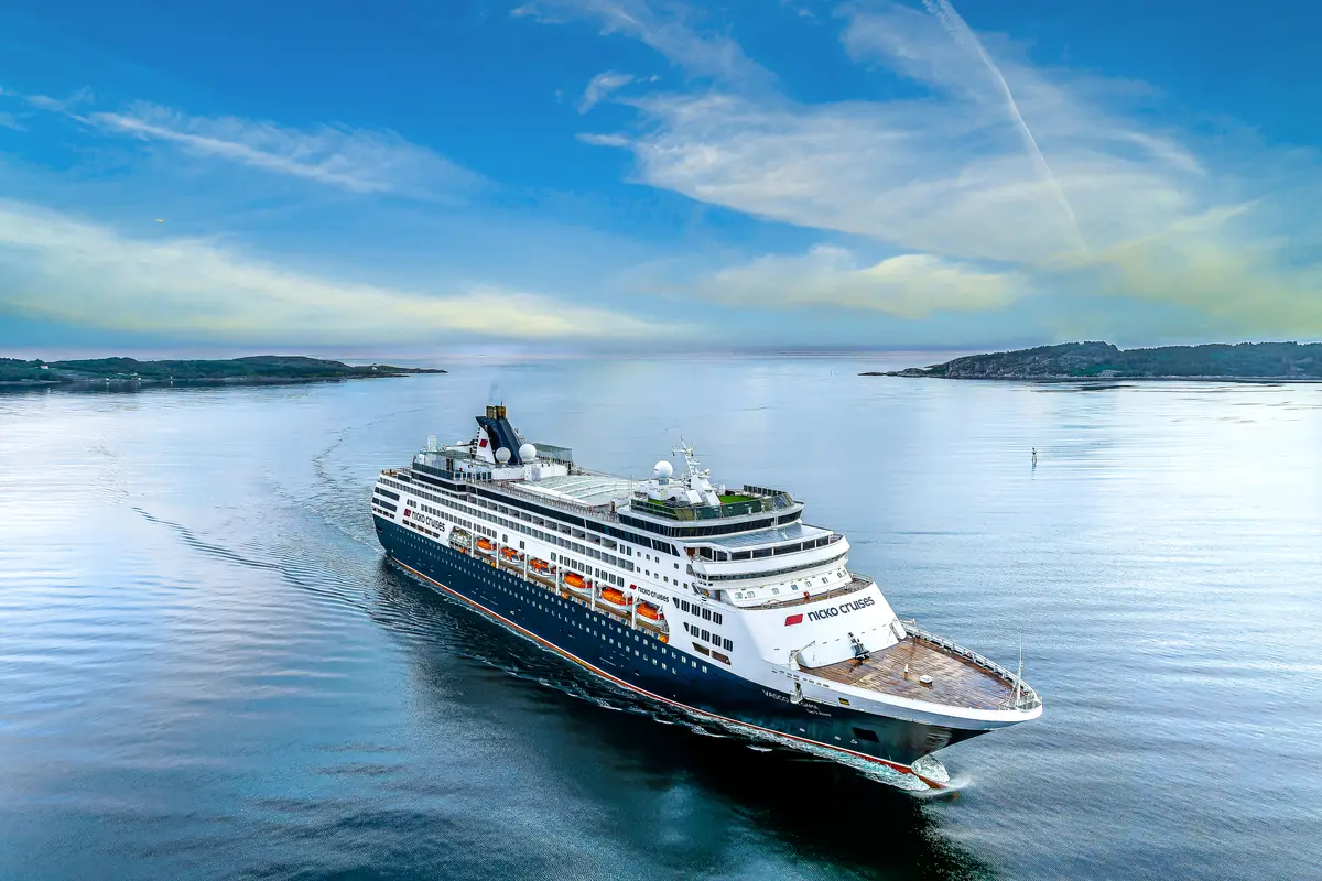 Vogelperspektive auf das dunkelblaue Kreuzfahrtschiff Vasco da Gama von Nicko Cruises, das auf ruhigem, blauem Wasser fährt. Im Hintergrund sind grüne Inseln und ein blauer Himmel mit Wolken zu sehen.