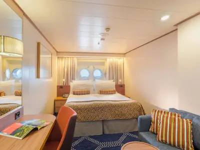 Innenansicht einer hellen Kabine des Flusskreuzfahrtschiffs Vasco da Gama mit Doppelbett, zwei Bullaugen, einem Sofa und Schreibtisch. Der Raum ist modern und wohnlich gestaltet.