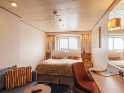 Helle Kabine auf der Vasco da Gama mit Doppelbett, Sofa, Schreibtisch und Fenster. Die moderne Einrichtung und der Ausblick auf eine Stadt sind deutlich erkennbar.