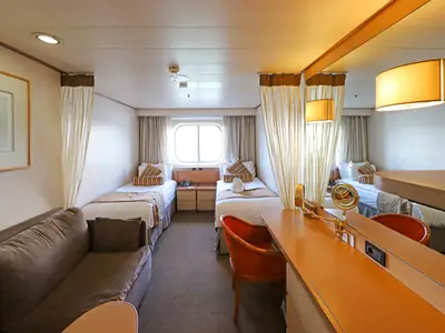 Innenansicht einer geräumigen Schiffskabine auf der Vasco da Gama mit zwei Einzelbetten, einem Sofa und einem Schreibtischbereich. Ein Bullauge bietet Tageslicht.