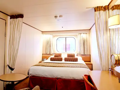 Innenansicht einer Kabine der Vasco da Gama. Im Fokus stehen das Doppelbett mit gestreiften Kissen, ein rundes Porthole-Fenster mit Vorhängen und ein Schreibtisch mit Spiegel.