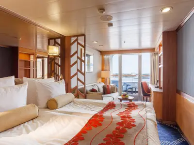 Helle Suite an Bord der Vasco da Gama. Im Vordergrund ein Bett, dahinter ein Wohnbereich mit Sofa und Balkon mit Meerblick. Holz und warme Farbtöne.