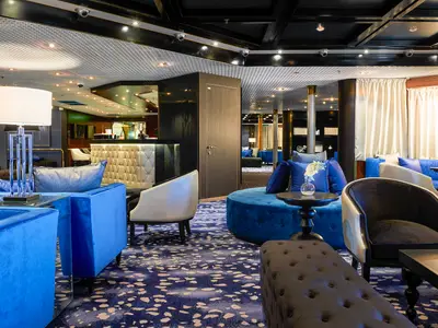 Eine helle und elegant eingerichtete Lounge auf dem Flusskreuzfahrtschiff Vasco da Gama. Zu sehen sind blaue Samtsofas, Tische, eine Bar und ein gemusterter Teppichboden.