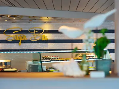 Ein Blick auf die "Sugar Bar" des Flusskreuzfahrtschiffs Vasco da Gama. Man sieht eine Theke mit Desserts, Gebäck und weiteren Speisen vor einer gestreiften Wand mit dem goldenen Schriftzug "Sugar Bar". Im Vordergrund ist eine Pflanze unscharf zu erkennen.