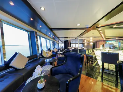 Innenansicht der Lounge des Flusskreuzfahrtschiffs Vasco da Gama. Die großen Fenster bieten weiten Ausblick. Zu sehen sind dunkle Sofas, blaue Sessel, kleine Tische und ein Bartresen mit Hockern.