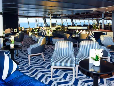 Die Lounge der Vasco da Gama zeigt blaue Sofas, gestreifte Sessel auf einem blau-weißen Teppich und Meerblick durch große Fenster. Im Hintergrund sind Bar und Bühne sichtbar.
