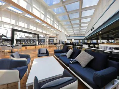 Einladende Lounge auf dem Kreuzfahrtschiff Vasco da Gama. Der helle Innenraum mit Glasdach bietet bequeme blaue Sitzgruppen, eine Bar und Blick auf einen Poolbereich mit Delfinskulptur.