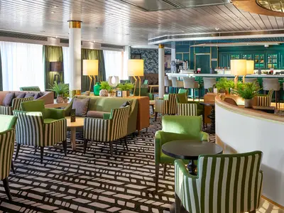 Der stilvolle Lounge- und Barbereich des Kreuzfahrtschiffs Vasco da Gama ist sichtbar. Grüne Sitzmöbel, Pflanzen und ein schwarz-weißer Teppich prägen den Raum.