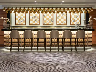 Moderne Bar mit sieben Barhockern, beleuchteter Theke mit nummerierten Sternkacheln. Darüber goldene Kugelleuchten und sechs Bilder Affen auf Schachbrettmuster. Holzrückwand.