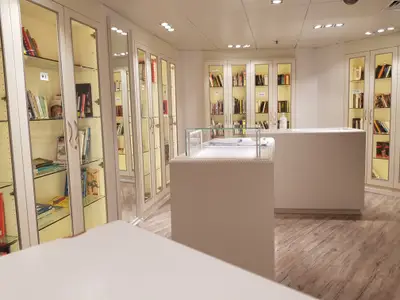Innenansicht der modernen Bibliothek des Flusskreuzfahrtschiffs Vasco da Gama. Zahlreiche beleuchtete Bücherregale mit Glastüren säumen die Wände, davor stehen weiße Präsentationstische.