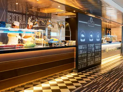 Zwei Köche servieren warme Speisen an einem modernen Buffet. Dunkle Wand mit Fleisch-Grafiken, Schachbrettmuster am Boden.