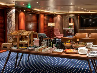 Buffet im Lounge-Bereich des Schiffs Vasco da Gama. Die Auslage bietet Backwaren, Müsli, Früchte und Getränke. Im Hintergrund sind gemütliche Sessel und Holzvertäfelungen sichtbar.