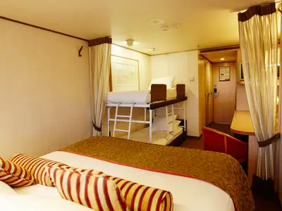 Helle Kabine auf der Vasco da Gama. Ein Doppelbett mit gestreiften Kissen im Vordergrund, dahinter Etagenbetten. Rechts ein Schreibtisch und die Kabinentür sind sichtbar.
