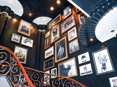Blick auf eine prächtige Treppe mit einem schmiedeeisernen Geländer. Die Wände sind mit einer Vielzahl gerahmter Schwarz-Weiß-Fotos und Kunstwerke dekoriert, die Decke zieren fächerförmige Elemente.