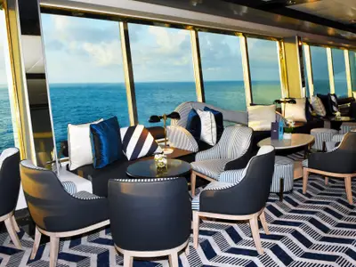 Lounge an Bord der Vasco da Gama mit großen Fenstern und Blick auf das Meer. Der Raum ist mit blauen Sofas, gestreiften Sesseln und einem blau-weißen Chevron-Teppich ausgestattet.