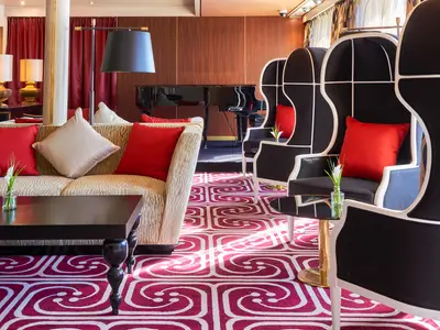 Eine stilvolle Lounge auf dem Flusskreuzfahrtschiff Vasco da Gama mit beigefarbenem Sofa, dunklem Couchtisch und mehreren schwarz-weißen Hochlehn-Sesseln. Im Hintergrund steht ein Flügel, der Boden ist mit einem auffälligen rot-weißen Teppich bedeckt.