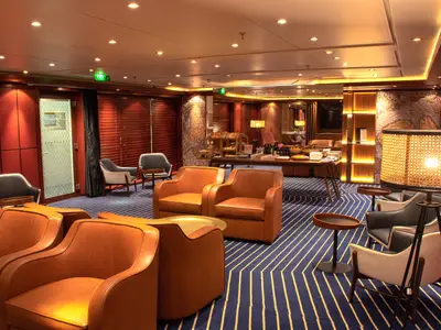 Elegante Lounge an Bord der Vasco da Gama. Das Bild zeigt mehrere braune Ledersessel, einen blauen Teppich mit auffälligem Muster und ein Buffet mit Gebäck. Der Raum ist warm beleuchtet, mit einer großen Stehlampe rechts.