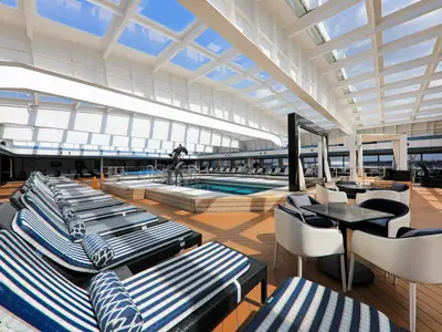 Blick auf den hellen Innenpoolbereich der Vasco da Gama. Man sieht blau-weiß gestreifte Liegen, einen Pool mit Delfinstatuen und moderne Sitzgruppen unter dem Glasdach.