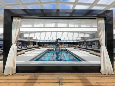 Der überdachte Poolbereich auf dem Kreuzfahrtschiff Vasco da Gama. Ein Pool mit Delfinbrunnen ist von Liegen gesäumt. Glasdach, Holzboden und Vorhänge.