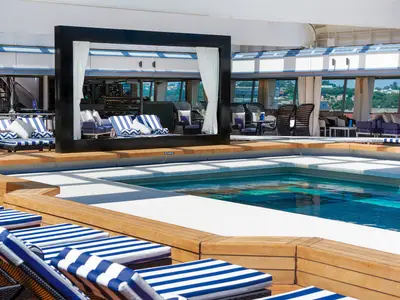 Das Pooldeck der Vasco da Gama mit blau-weiß gestreiften Liegen, einem Pool und Cabana. Dahinter ein verglaster Lounge-Bereich mit Sitzgruppen und Ausblick auf die Küste.
