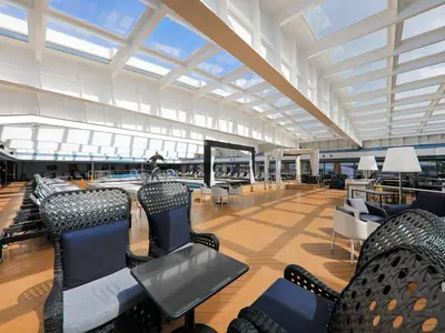 Geräumiges Sonnendeck der Vasco da Gama mit Loungemöbeln auf Holzboden. Ein großes Glasdach lässt viel Tageslicht auf den dahinterliegenden Poolbereich mit Liegestühlen fallen.