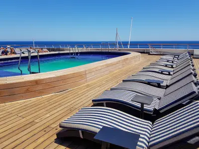 Auf dem Oberdeck der Vasco da Gama genießen Passagiere die Sonne am Pool. Reihen von blau-weiß gestreiften Sonnenliegen säumen das Holzdeck mit Weitblick auf das offene Meer.