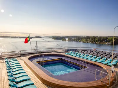 Das obere Deck der Vasco da Gama: Ein Pool umgeben von Liegestühlen. Portugiesische Flagge weht über einer Schärenlandschaft mit Inseln und Wasser. Sonniger Tag.