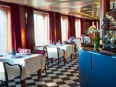 Im Restaurant der Vasco da Gama: gedeckte Tische, rote Vorhänge an Fenstern, Schachbrettboden. Rechts Theke mit Orchideen und großem schwarzen Vogelkäfig.