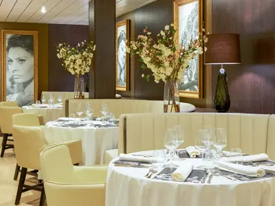 Elegantes Restaurant auf der Vasco da Gama. Helle Sitzmöbel und runde, gedeckte Tische prägen das Bild. Dunkle Holzwände sind mit Kunstwerken und großen Blumenvasen dekoriert.