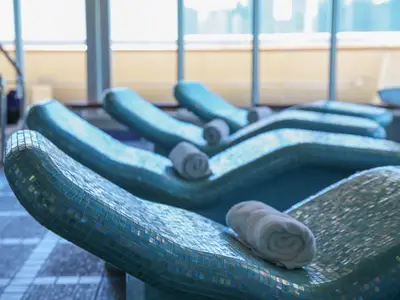 Nahaufnahme mehrerer blau-türkiser Mosaik-Relaxliegen mit gerollten weißen Handtüchern im Spa-Bereich der Vasco da Gama. Im Hintergrund sind große Fenster zu sehen.