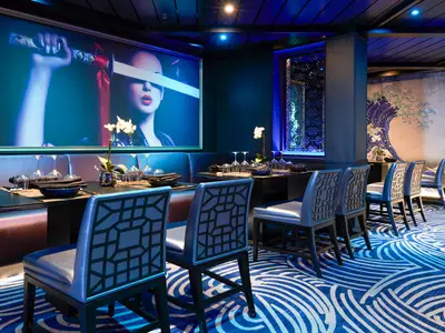 Innenansicht eines modern eingerichteten asiatischen Restaurants mit Tischen, Stühlen und einer Sitzbank. Eine große Kunstinstallation mit einer Frau und einem Katana ziert die Wand. Blaue Beleuchtung taucht den Raum in eine besondere Atmosphäre.