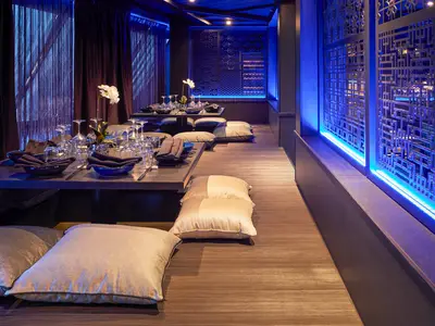 Innenansicht eines asiatisch inspirierten Spezialitätenrestaurants. Blaue LED-Beleuchtung akzentuiert die gemusterten Wandpaneele. Tische sind mit Geschirr und Servietten gedeckt, Sitzkissen liegen auf dem Boden für ein entspanntes Dining-Erlebnis.