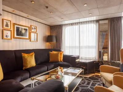 Luxuriöser Salon auf der Vasco da Gama: Dunkles Ecksofa, gelbe Kissen, Glastisch, Sessel und große Fenster mit Meerblick. Wände mit Kunstwerken.