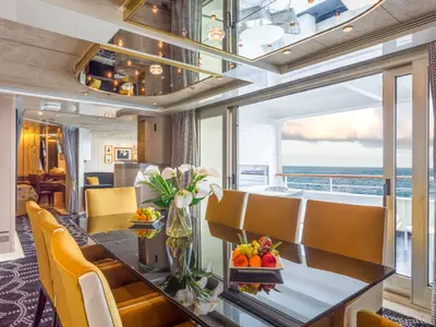 Die Suite der Vasco da Gama zeigt einen Essbereich mit dunklem Tisch, gelben Stühlen, Blumen und Obst. Glastüren führen zu einem Balkon mit weitem Meerblick unter bewölktem Himmel.