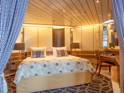 Kabinenansicht auf der Vasco da Gama: ein Doppelbett, Nachttische, Lampen und ein Schreibtischbereich. Links und rechts sind blau-weiß gemusterte Vorhänge. Warme Raumbeleuchtung.
