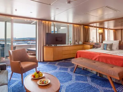 Helle Kabine auf dem Flusskreuzfahrtschiff Vasco da Gama. Großes Bett, Sitzbereich mit Sofa und Sessel, TV, Balkon mit Flussblick. Tisch mit Obst und Macarons.