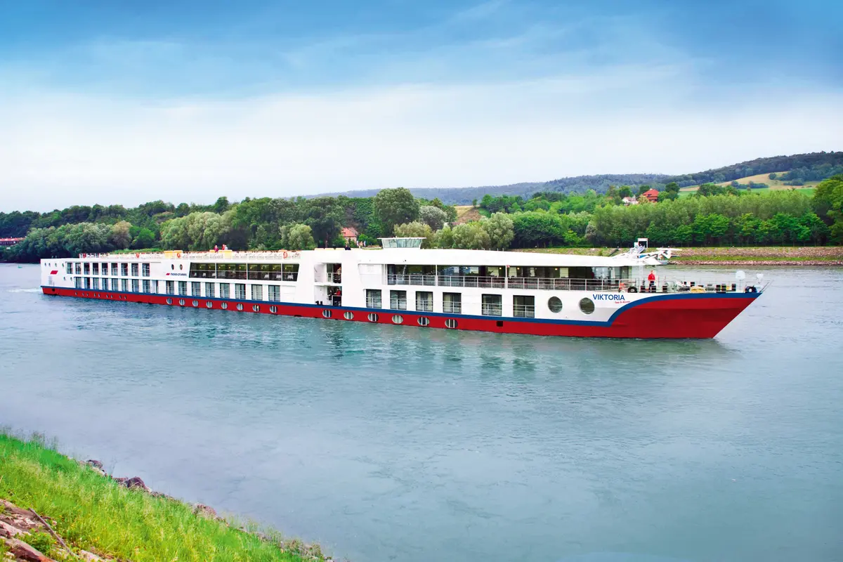 Das weiß-rote Flusskreuzfahrtschiff Viktoria auf einem Fluss. Grüne, bewaldete Uferlandschaft mit Hügeln und blauem Himmel im Hintergrund. Keine Personen.