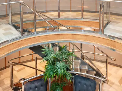 Flusskreuzfahrtschiff Viktoria: Treppenhaus aus Holz, Metall, Glas. Geschwungene Geländer. Pflanze und Sessel im Vordergrund. Heller Fliesenboden.