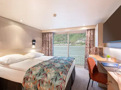 Moderne Kabine auf Flusskreuzfahrtschiff Viktoria. Doppelbett, Schreibtisch und Panoramafenster mit Blick auf Fluss und grüne Uferlandschaft sind zu sehen.