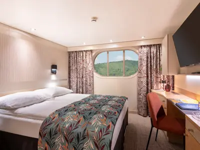 Eine helle Kabine an Bord des Flusskreuzfahrtschiffs Viktoria mit gemütlichem Bett, Schreibtisch und einem ovalen Fenster, das eine grüne Landschaft zeigt.