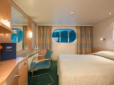 Blick in eine helle Schiffskabine der Viktoria mit einem Bett mit gemusterter Decke, Schreibtisch, Sessel und ovalem Fenster, das eine grüne Landschaft zeigt.