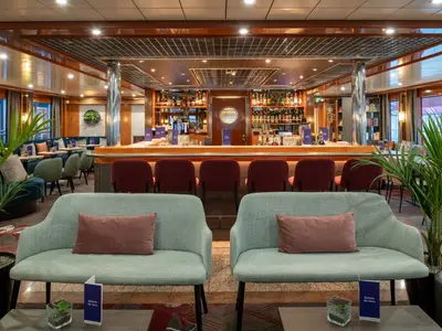 Die Bar des Flusskreuzfahrtschiffs Viktoria. Holztheke mit Barhockern, gefüllte Regale, helle Sofas, Tische und Pflanzen sind sichtbar. Seitlich weitere Sitzgruppen und Fenster.